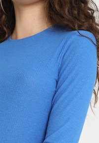 Primo piano di una persona che indossa una maglietta a maniche lunghe blu a coste, con capelli ricci castani e grandi orecchini a cerchio.