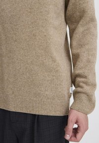 Maglione beige in lana con polsini e orlo a coste. Si nota il motivo a maglia strutturata e l'etichetta sul lato, abbinato a pantaloni scuri.