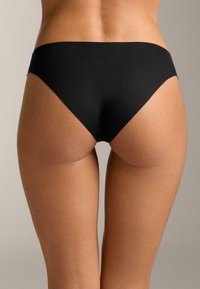 Žena v jednoduchých čiernych bikinových nohavičkách, zobrazená od dolnej časti chrbta po hornú časť stehien na neutrálnom pozadí.