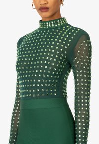 Abito verde con lunghe maniche trasparenti decorate con piccole strass quadrate. Design a collo alto con corpetto aderente e parte inferiore levigata.