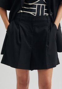 Černé upravené šortky s sklady vpředu, které mají boční kapsy a hladkou texturu, navržené pro uvolněný střih.
