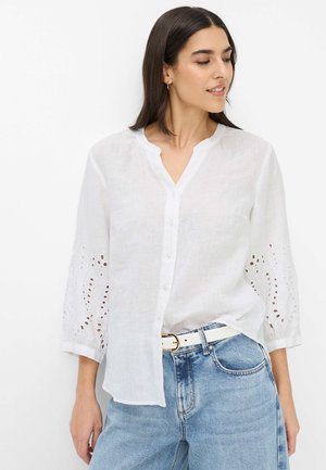 Frau trägt eine weiße, knöpfbare Bluse mit Ärmeln aus Lochspitze und hellblaue Jeans, blickt nach links vor einem schlichten weißen Hintergrund.