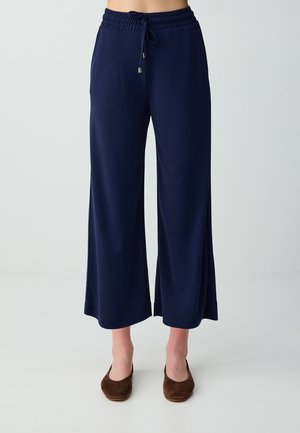 Persona che indossa pantaloni blu navy a gamba larga con vita elastica con coulisse e scarpe basse marroni, in piedi davanti a uno sfondo chiaro e semplice.