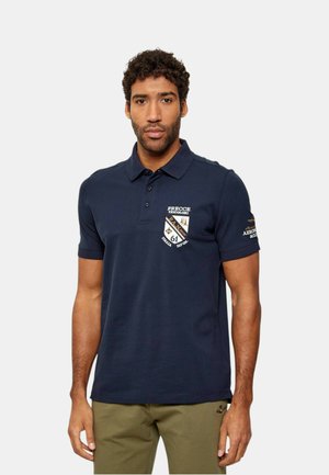 Uomo con capelli corti e ricci che indossa una polo blu navy con toppe ricamate e pantaloni verde oliva su uno sfondo chiaro e uniforme.