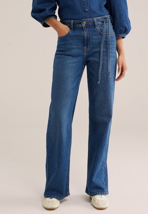 Femme portant un jean bleu taille haute à jambes larges avec une ceinture en denim assortie, un haut décontracté et des baskets blanches, debout avec une main dans la poche.