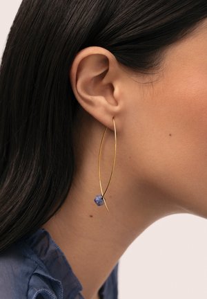 Primer plano de la oreja de una mujer con un pendiente de aro de alambre dorado largo que presenta una pequeña piedra azul, cabello oscuro y una blusa azul con volantes visibles.