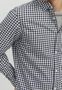 Camicia a quadri blu e bianchi, colletto con bottoni, maniche lunghe e morbido tessuto. I bottoni sono bianchi e posizionati lungo il pattino.