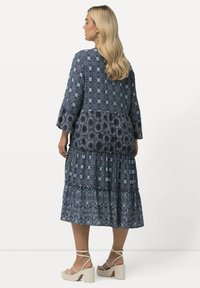 Vestido maxi con patrones en azul, que presenta volantes de tres niveles, mangas largas y un escote redondo. Combinado con sandalias de tacón cuadrado color beige.