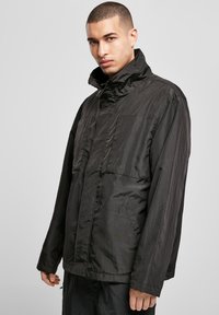 Juodas, lengvas švarkas su aukštu apykakle, priekiniu kišenėmis ir ilgomis rankovėmis. Lygi tekstūra ir minimalus dizainas su švelniu blizgesiu.