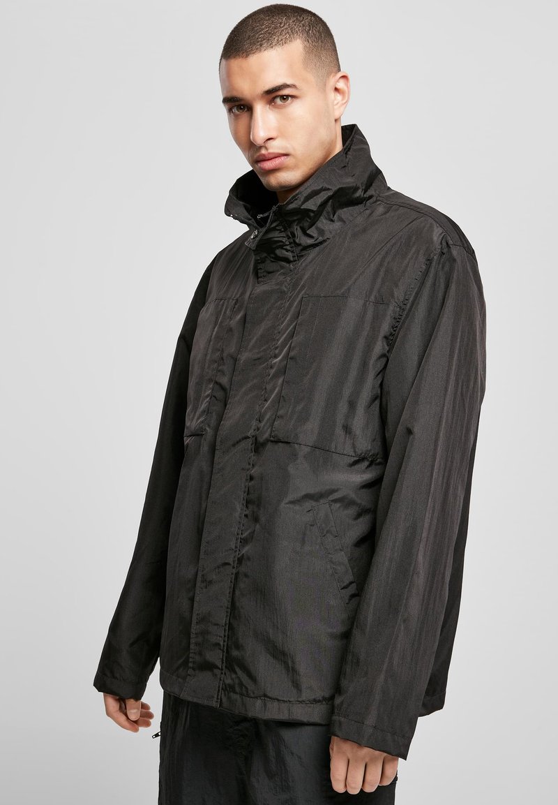 Juodas, lengvas švarkas su aukštu apykakle, priekiniu kišenėmis ir ilgomis rankovėmis. Lygi tekstūra ir minimalus dizainas su švelniu blizgesiu.