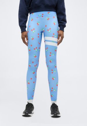 Leggings met lichtblauwe basis, rood aardbei- en wit bloemenprint, twee witte strepen op de rechterdij, gedragen met zwarte sneakers en navy sweatshirt.