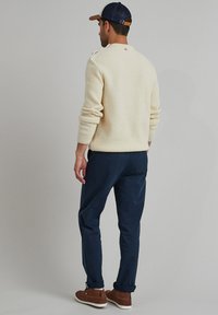 Cremfarbener Strickpullover mit strukturiertem Muster, marineblaue Hose und braune Wildlederschuhe. Die Mütze ist schwarz mit einem braunen Schild und Logodetail.