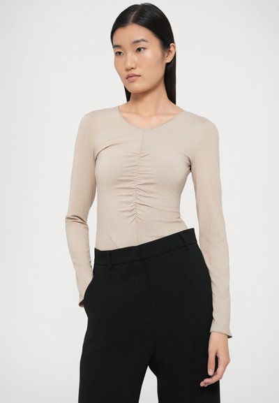 Hosszú ujjú, bézs színű felső kerek nyakkivágással és középen összegyűrt részlettel; magas derekú, fekete nadrággal párosítva. Sima anyagtextúra.