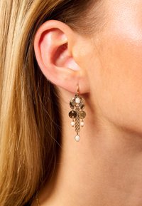 Boucles d'oreilles dorées avec des accents circulaires, des chaînes et des perles au fini texturé, suspendues en dessous du lobe de l'oreille.