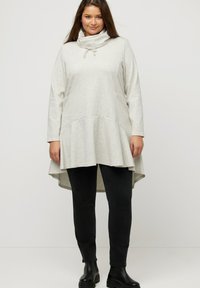 Túnica de manga larga gris claro con cuello tipo cowl y cordón, con parte inferior acampanada, realzada por bolsillos laterales, combinada con jeans skinny negros y botas.