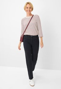 Chemise à rayures à manches longues blanche et bordeaux, accompagnée de pantalons foncés et de baskets blanches. Le modèle tient un sac bandoulière bordeaux.
