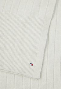 Kreminės spalvos raštuota megztinė skara, turinti mažą raudoną, baltą ir mėlyną logotipą pakraštyje. Minkšta tekstūra su šildančiu dizainu.