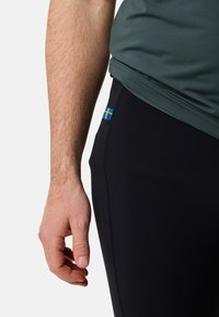 Pantalons de sport noirs ajustés avec une texture lisse, présentant un petit accent de drapeau suédois sur le côté et un t-shirt gris au-dessus.