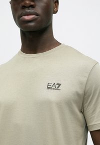 Helebeež värvi puuvillane T-särk ümarate kaelusega, millel on vasakul rinnal must EA7 Emporio Armani logo. Sujuv tekstuur, vormiv lõige.