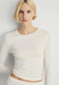 Top lung cu mâneci lungi, de culoare crem deschis, din material ribbed, cu gât rotund și siluetă ajustată. Textură netedă și design minimalistic.