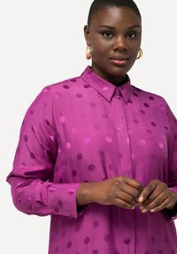 Chemise boutonnière violette avec un col, présentant des pois de tailles variées dans une teinte légèrement plus foncée. Texture lisse et manches longues.