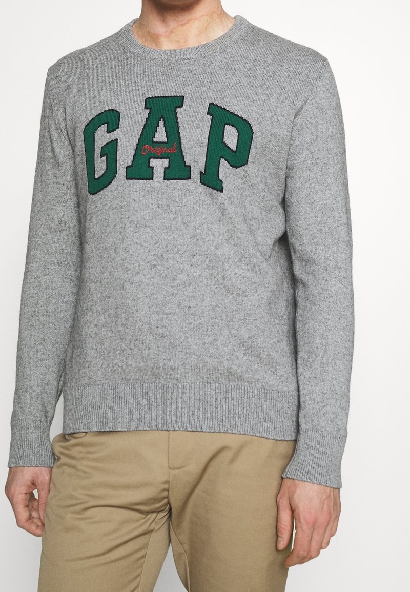 Persoon in een grijze gebreide trui met "GAP" in groene letters en beige broek, van nek tot halverwege de bovenbenen tegen een witte achtergrond.