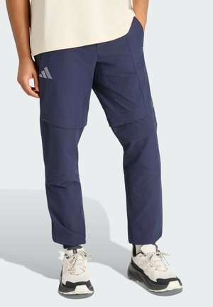 Osoba ubrana w granatowe spodnie zapinane na zamek z logo Adidas, białe i czarne trampki, stoi z jedną ręką w kieszeni.