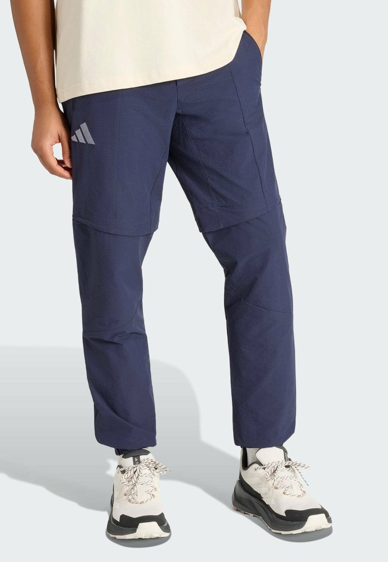 Persona con pantalones desmontables azul marino con el logo de Adidas, zapatillas blancas y negras, de pie con una mano en el bolsillo.