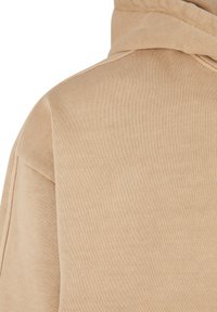 Beige kapucnis felső, pamut anyagból, puha tapintással, látható varrásokkal a varratok mentén, és egy nagy kapucnival a nyaknál.