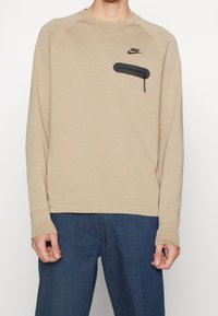 Beżowy długi rękaw Nike z kieszenią na zamek błyskawiczny na klatce piersiowej, noszony z niebieskimi teksturowanymi spodniami.