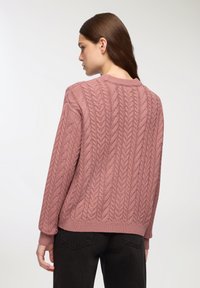 Sweter w warkocz w odcieniu przybrudzonego różu, z prążkowanymi mankietami i dołem. Tekstura jest gruba z wypukłymi wzorami, co zapewnia przytulny, miękki wygląd.