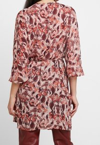 Blusa ligera de longitud media en beige con intrincados patrones de paisley en rojo y marrón. Presenta mangas de tres cuartos y un lazo en la cintura.