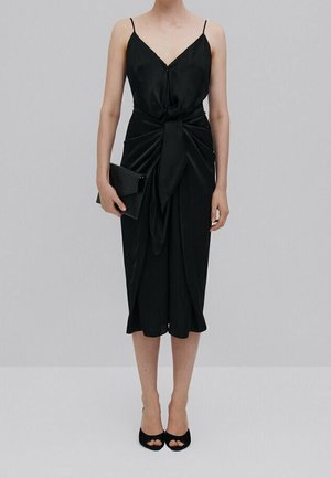 Vrouw in een zwart midi-jurkje met spaghetti-bandjes en een voorzijde met strikdetail, die een zwarte clutch vasthoudt en zwarte open-teenhakken draagt.