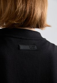 Crna dukserica s teksturiranim materijalom, okruglim izrezom i mat crnom etiketom s utisnutim logotipom na leđima.
