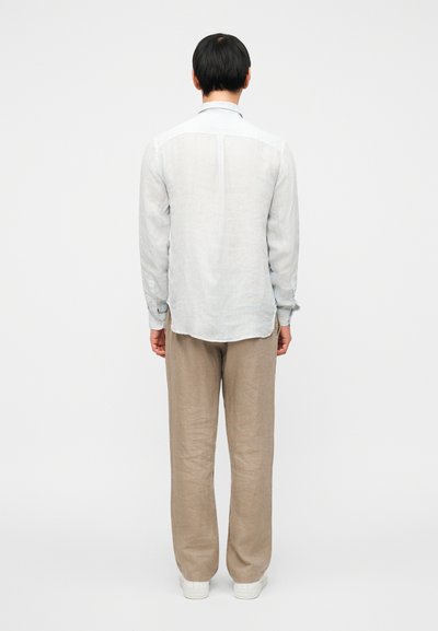 Ανοιχτό γαλάζιο λινό πουκάμισο μακρυμάνικο με απλή πλάτη και μανσέτες με κουμπιά, φορεμένο με μπεζ λινό παντελόνι και λευκά αθλητικά παπούτσια.