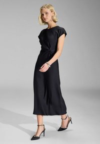 Sort satin kjole med korte ærmer, med et twist-detalje ved taljen. Bært med sorte høje hæle og et sølvarmbånd. Glat tekstur.