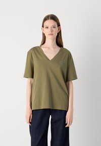 Olivově zelené bavlněné tričko s výstřihem do V, krátkými rukávy a malým vyšitým logem Lacoste na hrudi. Model má na sobě tmavé džíny.