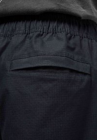 Dunkle, strukturierte Shorts mit einem elastischen Bund und einer einzigen Gesäßtasche mit flacher Verschlussschließe. Der Stoff hat ein Karomuster.
