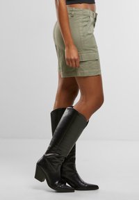 Kobieta ubrana w oliwkowozielone krótkie spodenki typu cargo i czarne, skórzane kozaki za kolano, stojąca na tle gładkiego, jasnego tła.