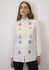 Bela bluzica z barvitim cvetličnim vezenjem v roza, rumeni in modri barvi. Ima visok ovratnik, zapiranje na gumb in strukturiran rob.