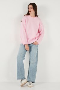 Mujer de pie contra un fondo blanco, vistiendo un suéter rosa grande, jeans de pierna ancha azul claro y zapatillas blancas, mirando de lado.