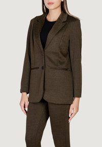 Blazer marrón de espiga con collar de pico, cierre de un solo botón y dos bolsillos frontales. La tela texturizada ofrece un aspecto a medida.