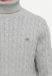 Hellgrauer, kabelgenoppter Rollkragenpullover mit kleinem, gesticktem Wappenemblem auf der Brust.