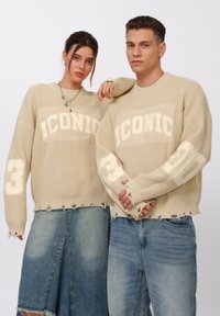 Beige knit sweater featuring "ICONIC" text, frayed hem, and numbers on sleeves. Worn with distressed denim skirts and jeans.
