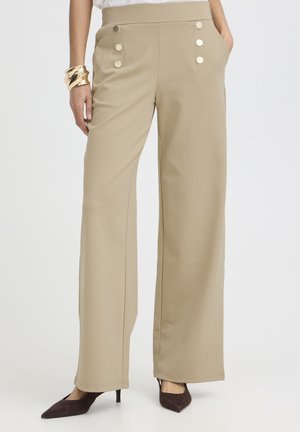 Pantaloni beige a vita alta a gamba larga con bottoni decorativi bianchi, indossati con tacchi scuri a punta e un braccialetto d'oro al polso sinistro.