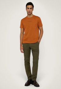 Camiseta de algodón naranja con cuello redondo, con un sutil logo de "Trussardi". Combinada con pantalones chinos verde oliva y mocasines marrón oscuro.