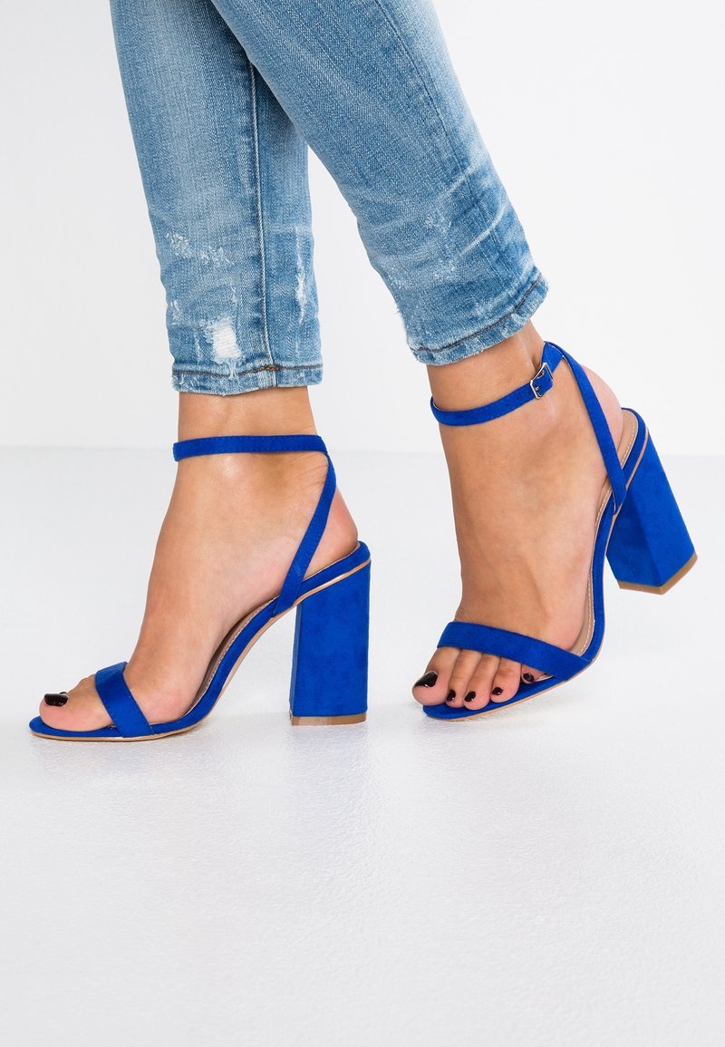 Kuningan biru royal dengan hak blok yang ramping, menampilkan tali lebar di atas jari kaki dan pergelangan kaki. Terbuat dari bahan suede yang lembut.