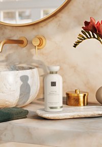Bouteille blanche de "Shampooing Récupérateur" avec des accents verts, posée sur un plateau en marbre beige texturé, entourée d'une serviette verte et de décorations dorées.