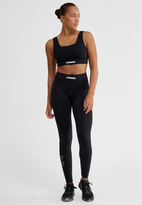 Schwarzes Sport-Crop-Top und hoch taillierte Leggings mit Hummel-Logo. Die Leggings haben ein Chevron-Muster am Saum. Kombiniert mit schwarzen Sneakern.