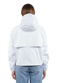 Giacca leggera bianca con cappuccio, caratterizzata da un design ampio e una patta sul retro. Il tessuto appare liscio ed è privo di decorazioni. Indossata sopra dei jeans.